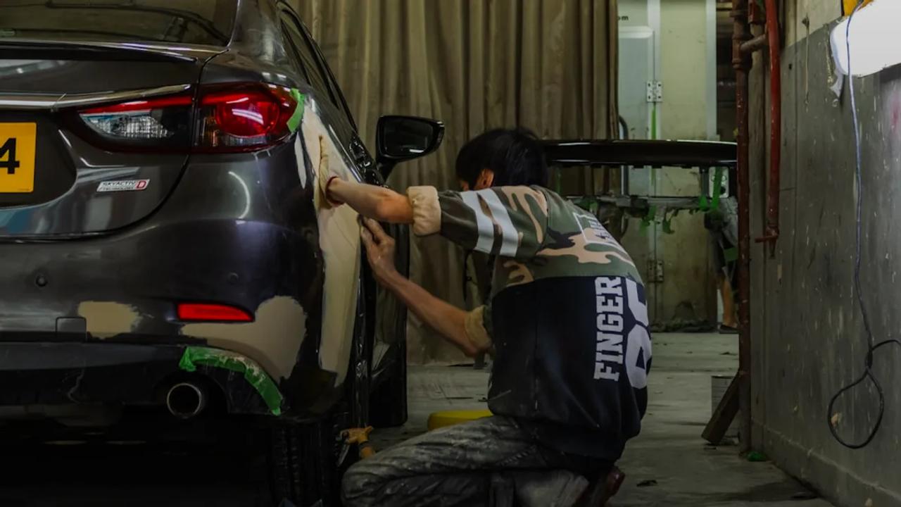 A person sanding the rear panel of a grey car in a workshop.