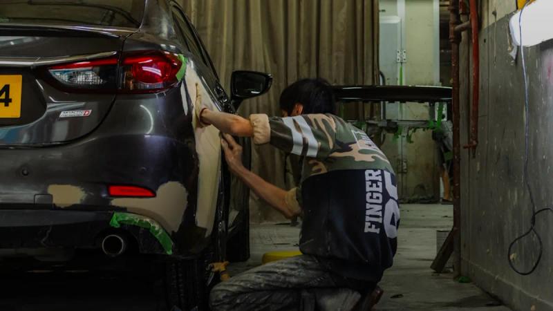 A person sanding the rear panel of a grey car in a workshop.