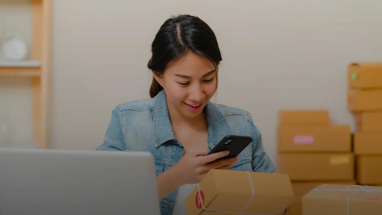Woman checking package with phone near laptop and boxes