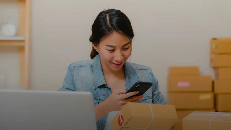 Woman checking package with phone near laptop and boxes